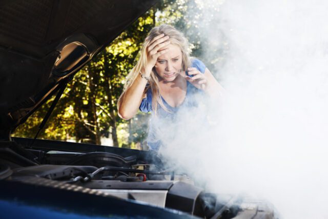 Cette jeune femme désemparée, immobilisée après une panne de voiture, regarde le moteur fumant, la main sur la tête, grimaçant entre confusion horrifiée et frustration.