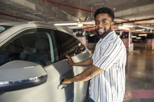 Homme noir souriant ouvrant la porte de sa voiture et regardant la caméra dans un parking souterrain public.