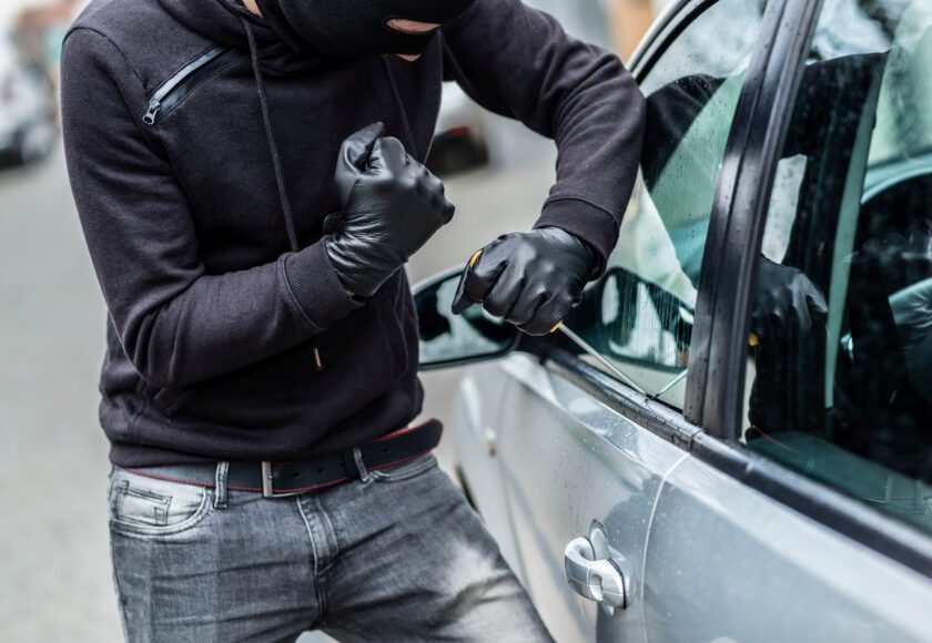 Un homme vêtu de noir, cagoule sur la tête, tente de s’introduire dans une voiture à l’aide d’un tournevis. Concept de vol et de tentative de cambriolage de voiture.